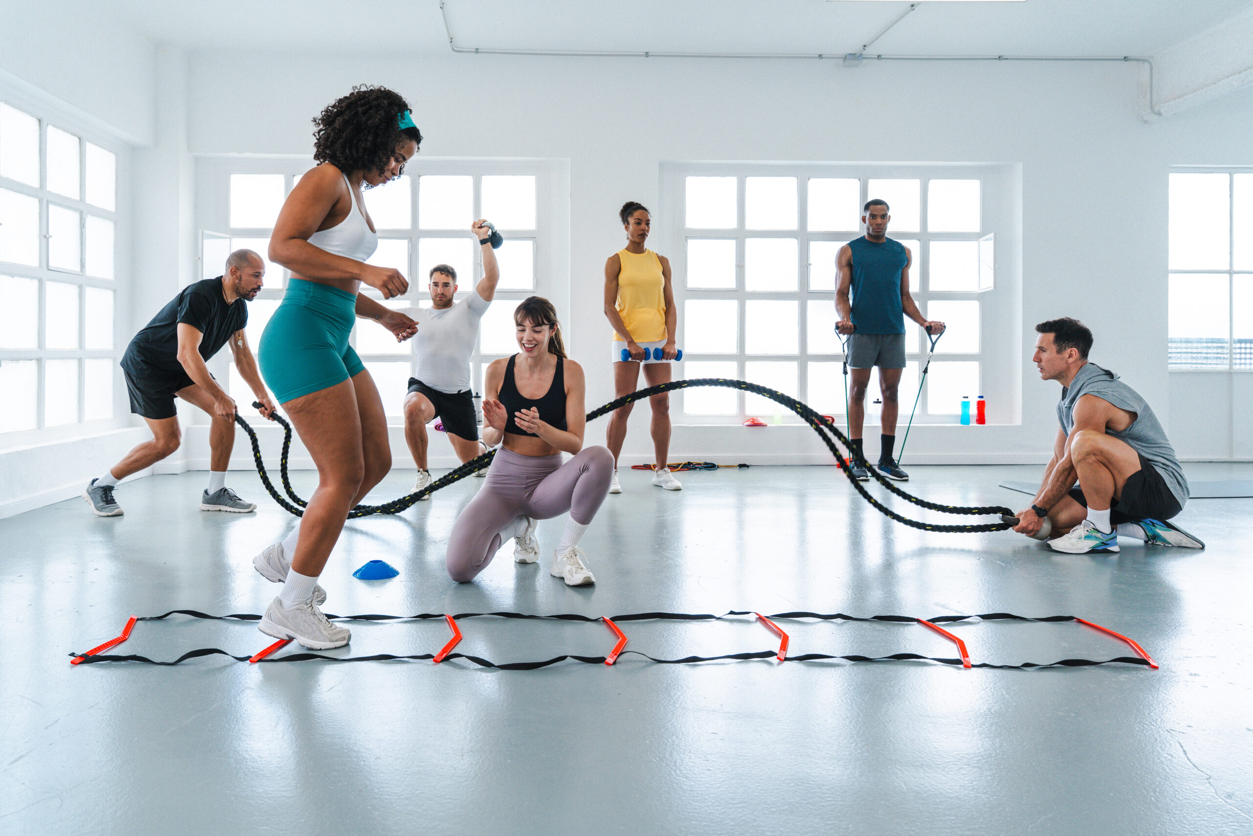 Multiethnic group of sportive people doing functional workout in training class gym diverse athletes exercising with battle ropes and agility ladder under personal trainer's guidance