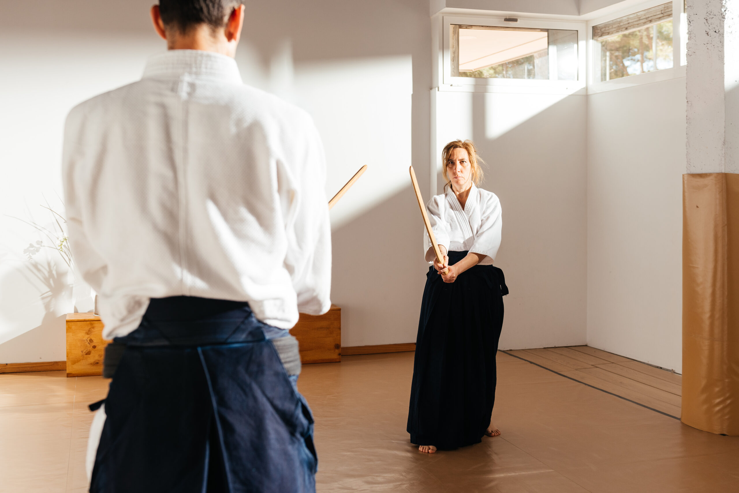 Female martial artist, in traditional attire, focus intently on male partner as they practice with wooden swords in a sunlit dojo.